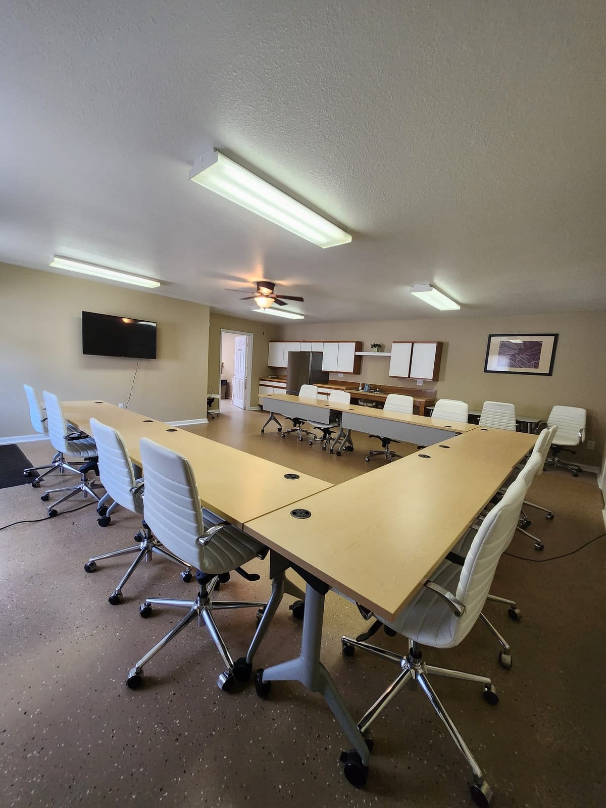 Another view of the community room at Prince Frederick Senior, showing a wall-mounted television, maple tables, white rolling chairs, and the kitchenette beyond