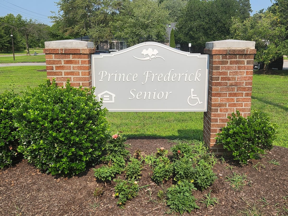 The Prince Frederick Senior entrance sign, a brick monument sign reading Prince Frederick Senior with fair-housing and accessibility symbols, framed by shrubs and flowers